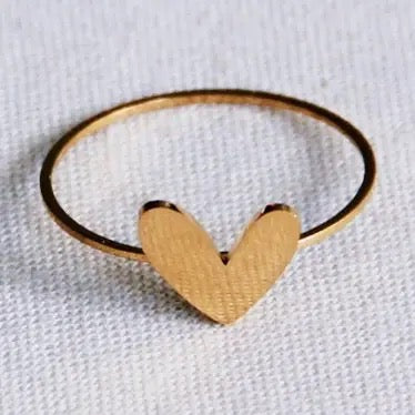 Minimalistischer, goldfarbener Edelstahlring von Bazou mit einem kleinen, flachen Herz-Motiv auf einem hellen, strukturierten Stoffhintergrund.