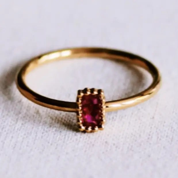 Filigraner, goldfarbener Edelstahlring von Bazou mit einem kleinen, rechteckigen, facettierten pinken Zirkonia-Schmuckstein in einer feinen Goldfassung auf hellem Stoffhintergrund.