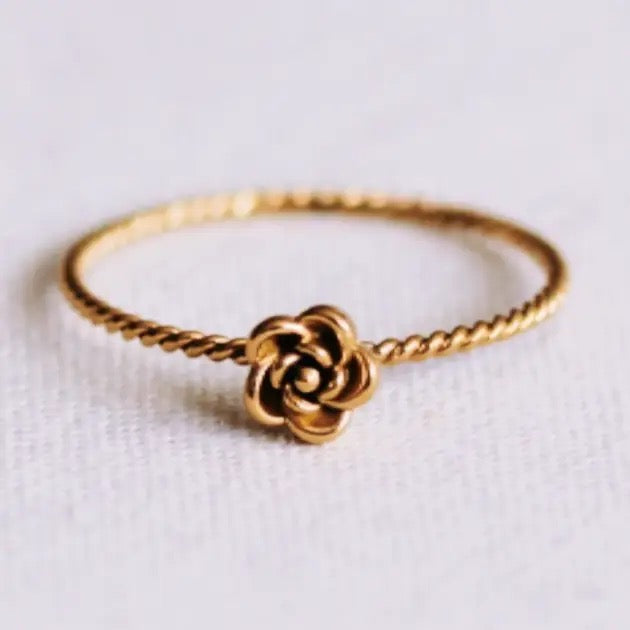 Filigraner, goldfarbener Edelstahlring von Bazou mit einer kleinen, detaillierten Rose in der Mitte und einer fein gedrehten Ringschiene auf hellem Stoffhintergrund.