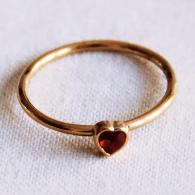 Filigraner, goldfarbener Edelstahlring von Bazou mit einem kleinen, glänzenden roten Zirkonia-Stein in Herzform auf einem hellen, strukturierten Stoffhintergrund.