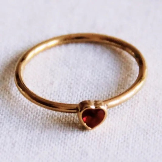 Filigraner, goldfarbener Edelstahlring von Bazou mit einem kleinen, glänzenden roten Zirkonia-Stein in Herzform auf einem hellen, strukturierten Stoffhintergrund.