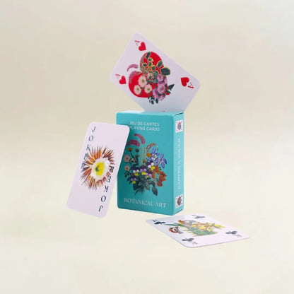 Eine kreative und dynamische Produktpräsentation vor einem hellen, neutralen Hintergrund. Die türkisfarbene „Botanical Art“-Box steht aufrecht. Auf ihrer Oberkante balanciert schräg das Herz-As, das ein großes rotes Herz voller Blumen zeigt. Links daneben schwebt eine Joker-Karte mit einer großen, sonnenartigen Blüte, während im Vordergrund eine Kreuz-König-Karte flach auf dem Boden liegt.