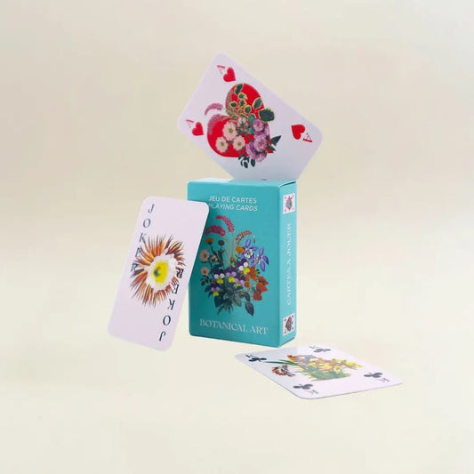 Eine kreative und dynamische Produktpräsentation vor einem hellen, neutralen Hintergrund. Die türkisfarbene „Botanical Art“-Box steht aufrecht. Auf ihrer Oberkante balanciert schräg das Herz-As, das ein großes rotes Herz voller Blumen zeigt. Links daneben schwebt eine Joker-Karte mit einer großen, sonnenartigen Blüte, während im Vordergrund eine Kreuz-König-Karte flach auf dem Boden liegt.