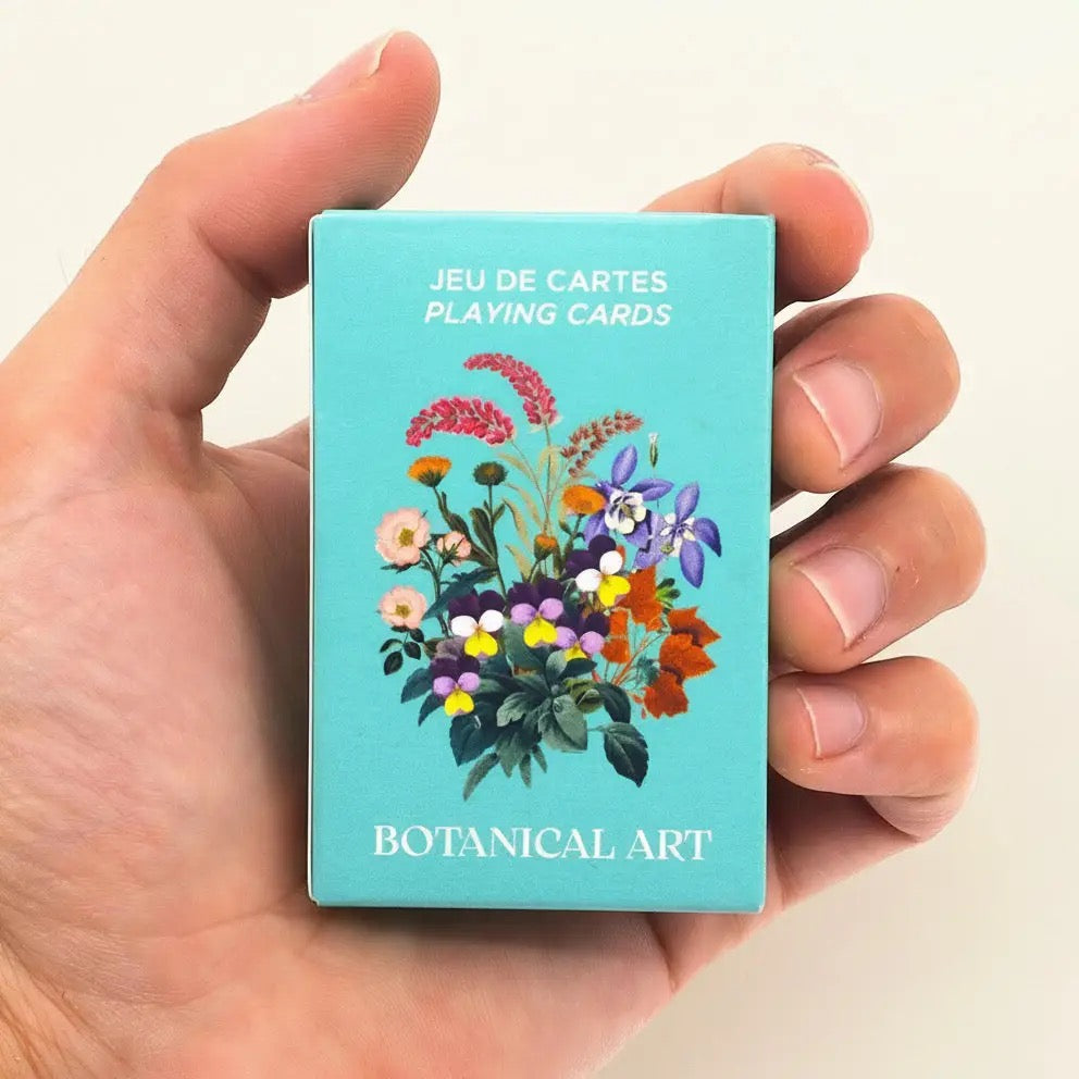 Eine geöffnete Hand hält die kompakte, geschlossene Produktbox des „Botanical Art“ Kartenspiels, wodurch die handliche Größe verdeutlicht wird. Die Box ist in einem kräftigen Türkis gestaltet. Auf der Vorderseite prangt eine feine Illustration eines bunten Wildblumenstraußes mit Stiefmütterchen und Vergissmeinnicht. Oben steht der Text „JEU DE CARTES / PLAYING CARDS“ und unten der Titel „BOTANICAL ART“.
