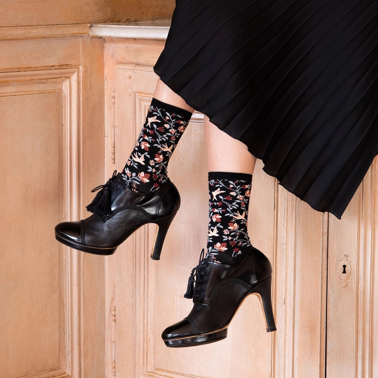 Detailaufnahme der schwarzen Socken mit Vogel- und Blumenmuster, getragen in Kombination mit schwarzen Pumps und einem schwarzen Faltenrock.