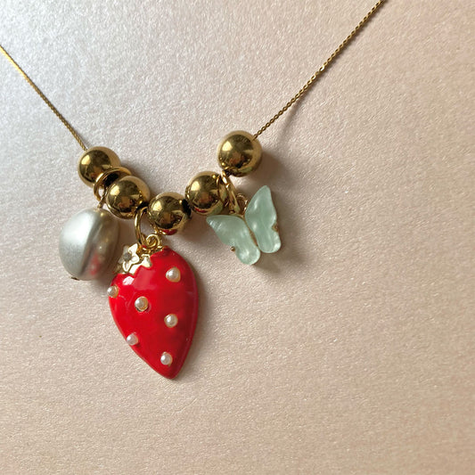 Halskette mit goldenen Kugeln, rotem Erdbeeranhänger mit weißen Punkten, weißem Perlenanhänger und hellgrünem Schmetterlingsanhänger.