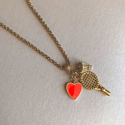 Goldene Halskette aus Edelstahl mit drei filigranen Anhängern: einem detaillierten Badminton-Schläger, einem kleinen Federball mit weißer Spitze und einem leuchtend orangefarbenen Herz-Charm, fotografiert auf einem hellen Hintergrund.