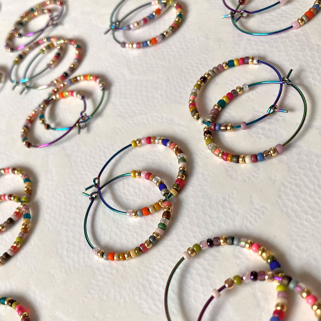 Bunte Perlen-Creolen in verschiedenen Farben auf einem hellen Untergrund. Die handgefertigten Ohrringe zeigen eine bunte Mischung aus Glasperlen und metallischen Akzenten an regenbogenfarbenen Metallreifen.