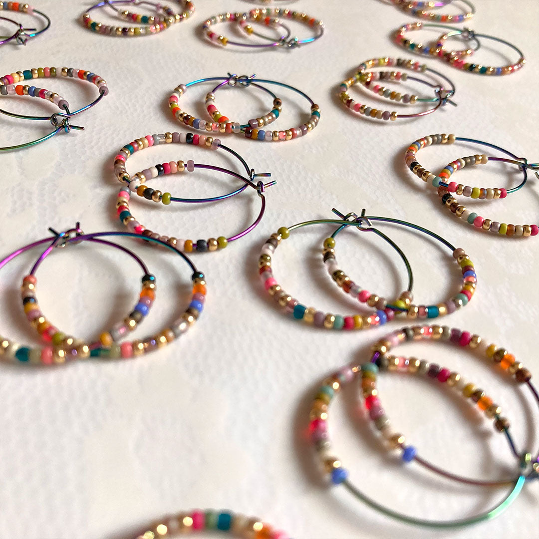 Bunte Perlen-Creolen in verschiedenen Farben auf einem hellen Untergrund. Die handgefertigten Ohrringe zeigen eine bunte Mischung aus Glasperlen und metallischen Akzenten an regenbogenfarbenen Metallreifen.