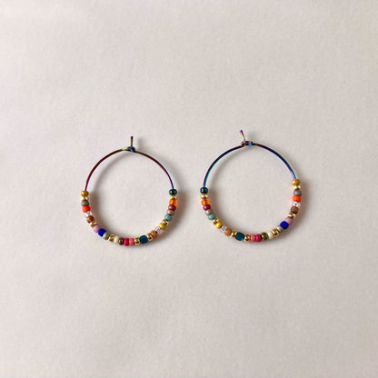 Bunte Perlen-Creolen in verschiedenen Farben auf einem hellen Untergrund. Die handgefertigten Ohrringe zeigen eine bunte Mischung aus Glasperlen und metallischen Akzenten an regenbogenfarbenen Metallreifen.