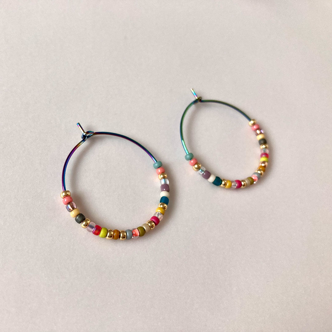 Bunte Perlen-Creolen in verschiedenen Farben auf einem hellen Untergrund. Die handgefertigten Ohrringe zeigen eine bunte Mischung aus Glasperlen und metallischen Akzenten an regenbogenfarbenen Metallreifen.
