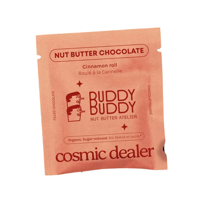 Cosmic Dealer x Buddy Buddy Nut Butter Chocolate Cinnamon Roll Verpackung auf lachsfarbenem Hintergrund, umgeben von Zimtstangen, Haselnüssen und Mandeln.