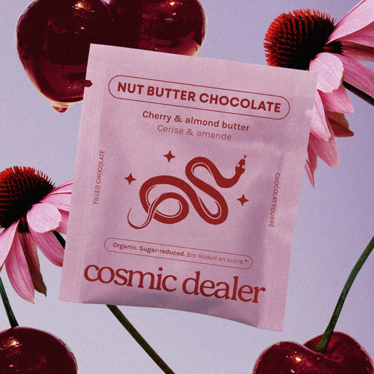 Hellviolette Verpackung der Cosmic Dealer Schokolade „Cherry & almond butter“ im Zentrum, umgeben von schwebenden, glänzenden Kirschen und rosafarbenen Sonnenhut-Blüten (Echinacea) auf lila Hintergrund.