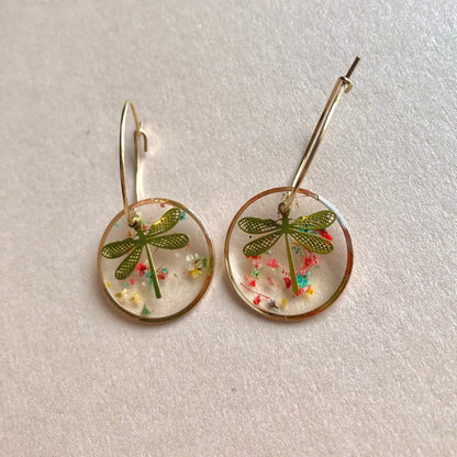 Goldene runde Ohrhänger mit durchsichtigen Resin-Scheiben, in denen eine grüne Libelle und bunte kleine Blüten eingeschlossen sind.