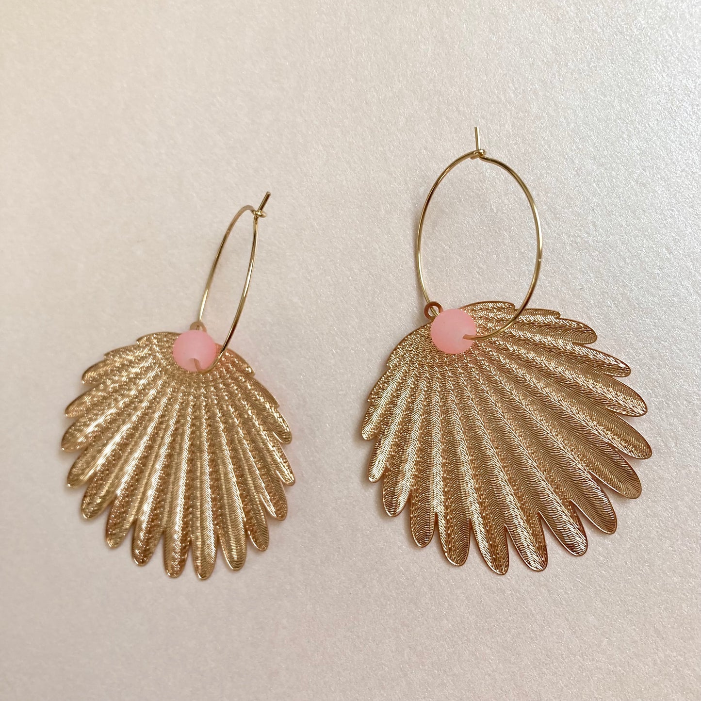 Goldfarbene Ohrringe in Federform mit feiner Struktur und rosafarbenem Perlenakzent an dünnem Bügel.