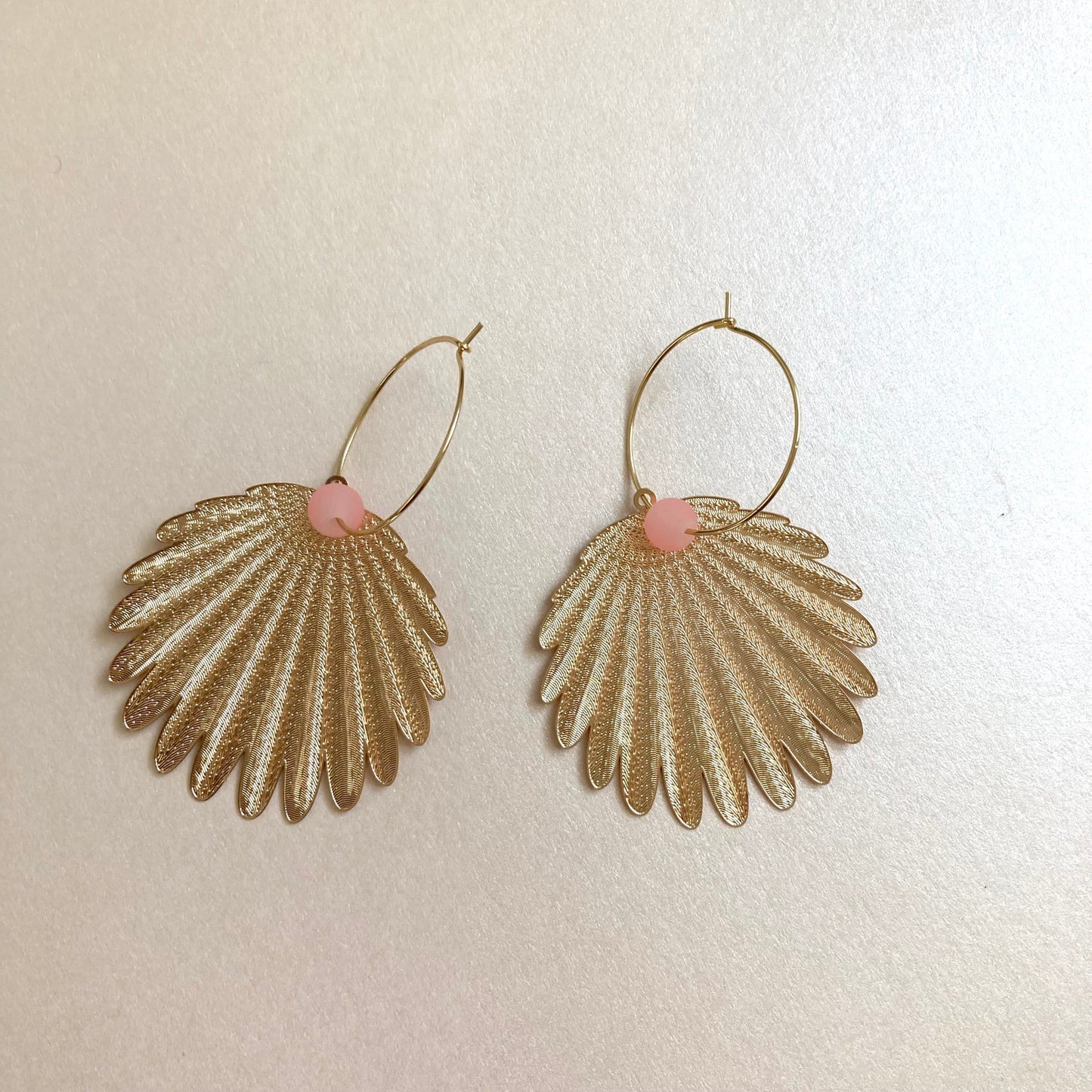 Goldfarbene Ohrringe in Federform mit feiner Struktur und rosafarbenem Perlenakzent an dünnem Bügel.