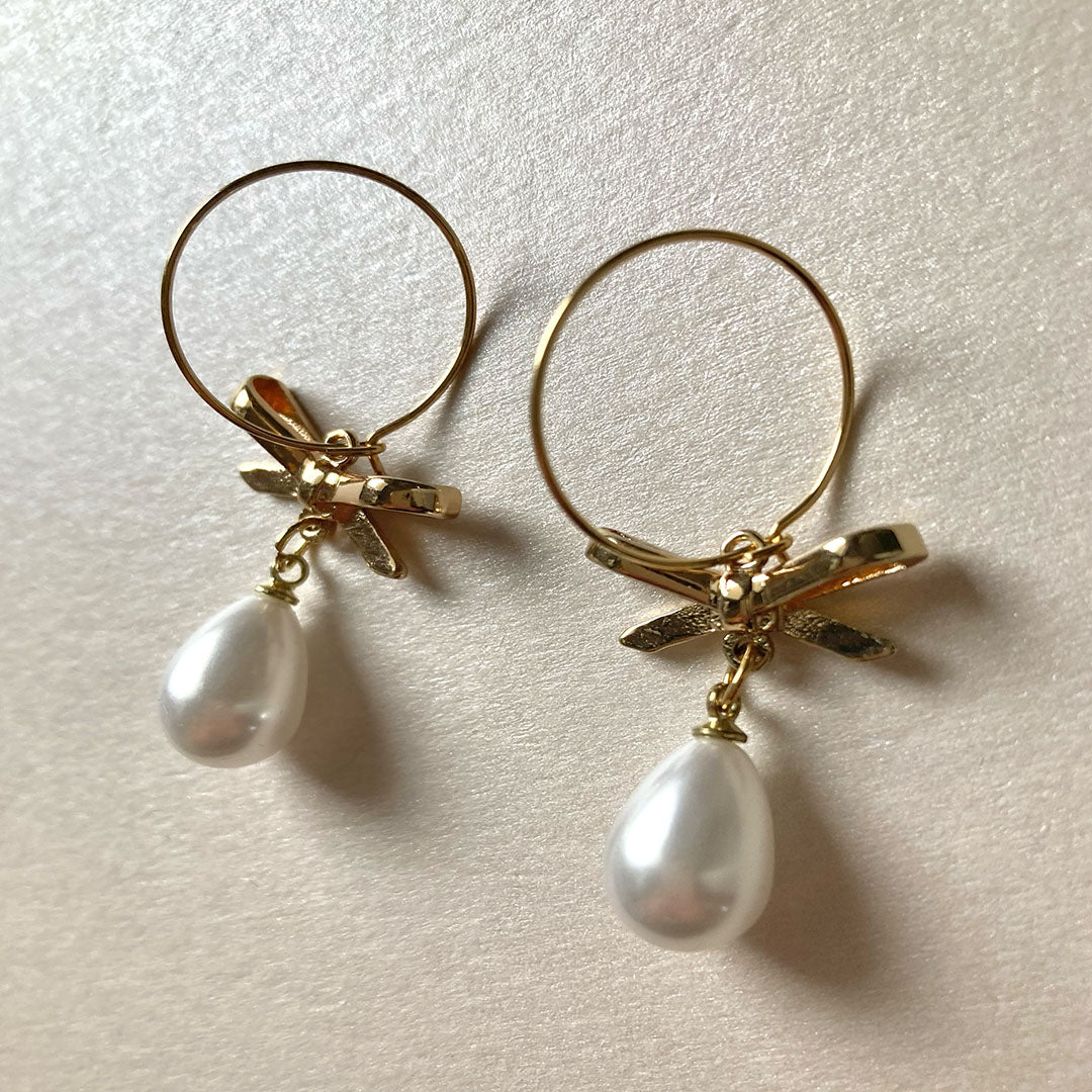 Ein Paar goldene Creolen mit einem romantischen Schleifen-Detail in der Mitte. An der Unterseite der Schleife hängt jeweils eine weiße, tropfenförmige Perle mit sanftem Glanz.