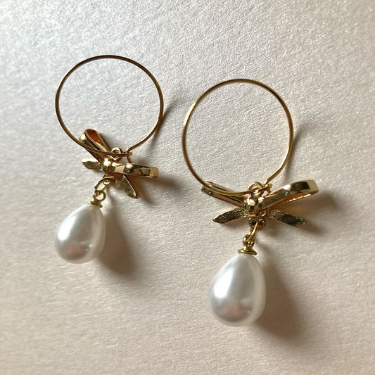 Ein Paar goldene Creolen mit einem romantischen Schleifen-Detail in der Mitte. An der Unterseite der Schleife hängt jeweils eine weiße, tropfenförmige Perle mit sanftem Glanz.