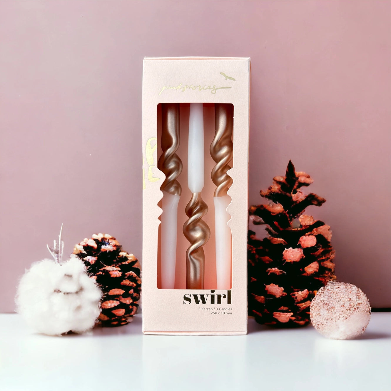 Drei gedrehte Kerzen in einer hellrosa Verpackung mit Sichtfenster, umgeben von zwei mit rosa Farbe besprühten Kiefernzapfen und einer weißen Dekoration.