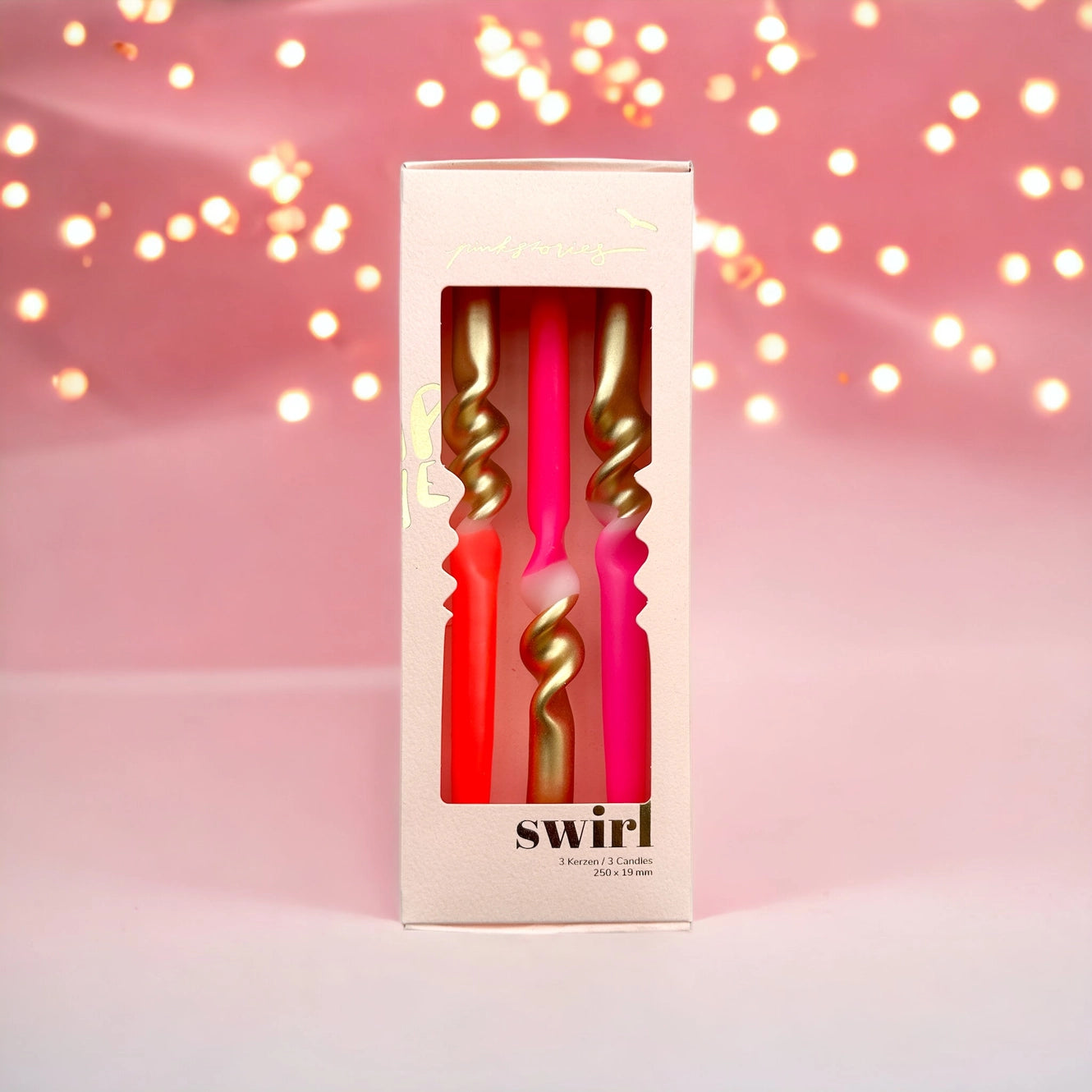 Verpackung mit drei Kerzen, zwei pink und eine rot, jeweils mit goldfarbenem Spiraldesign, vor rosa Hintergrund mit Lichterkette.
