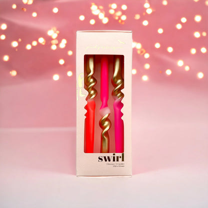 Verpackung mit drei Kerzen, zwei pink und eine rot, jeweils mit goldfarbenem Spiraldesign, vor rosa Hintergrund mit Lichterkette.