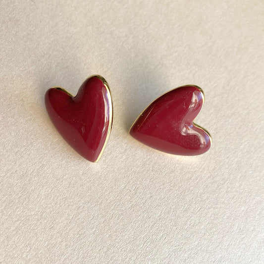 Zwei weinrote Emaille-Herzohrringe mit goldenem Rand, kunstvoll arrangiert auf einer beigen Textur-Oberfläche.