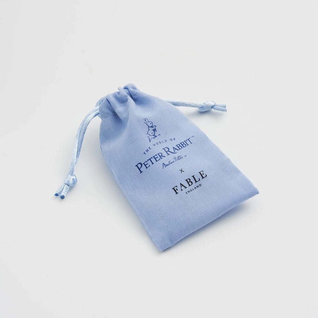 Ein kleiner hellblauer Stoffbeutel mit Kordelzug, bedruckt mit dem Logo „The World of Peter Rabbit x Fable England“.