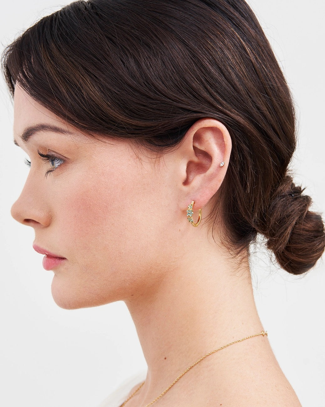 Seitenprofil einer Frau mit dunklem, hochgestecktem Haar, die eine goldene Fable England Creole mit blau emaillierten Vergissmeinnicht-Blüten trägt. Der Schmuck schmiegt sich dezent an das Ohrläppchen an.