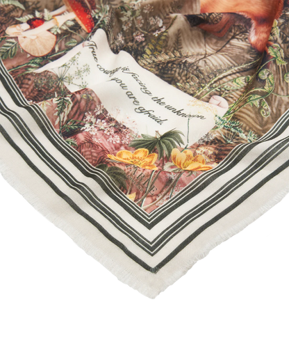 Nahaufnahme einer Ecke des Fable England Schals mit feinen Fransen, gestreifter Bordüre und dem Teil des Zitats „facing the unknown when you are afraid.“ neben einer gelben Blume.