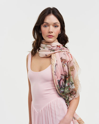 Frau mit dunklen Haaren trägt einen großen, rosafarbenen Fable England Schal mit Naturmotiven locker über die Schultern zu einem passenden rosa Kleid.