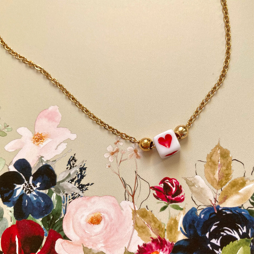 Zarte goldene Gliederkette mit einem kleinen weißen Würfel in der Mitte, auf dem ein rotes Herz abgebildet ist.