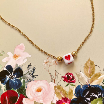 Zarte goldene Gliederkette mit einem kleinen weißen Würfel in der Mitte, auf dem ein rotes Herz abgebildet ist.