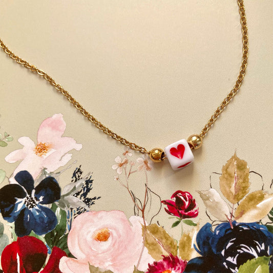 Zarte goldene Gliederkette mit einem kleinen weißen Würfel in der Mitte, auf dem ein rotes Herz abgebildet ist.