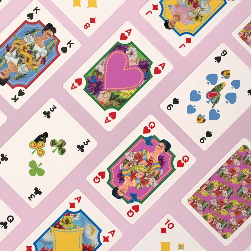 Eine dynamische Nahaufnahme verschiedener Spielkarten, die schräg über einen hellrosa Hintergrund verteilt sind. Zu sehen sind unter anderem das As mit einem großen rosa Herz, umgeben von floralen Mustern, ein König in Blau-Rot, eine Dame in Grün-Magenta sowie Zahlenkarten mit kunstvoll verzierten Symbolen. Unten rechts ist die gemusterte Rückseite der Karten mit einem dichten, floralen Frida-Kahlo-Motiv sichtbar.