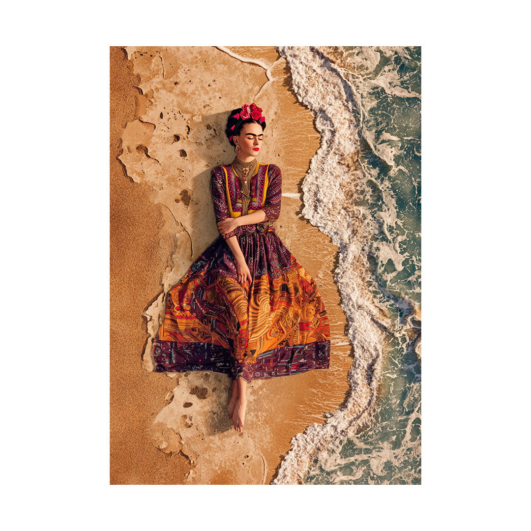 Vogelperspektive einer Frau im Frida-Kahlo-Look mit geschlossenem Blick, die barfuß in einem aufwendig gemusterten Kleid am Sandstrand liegt, während sanfte weiße Meereswellen sie umspülen.