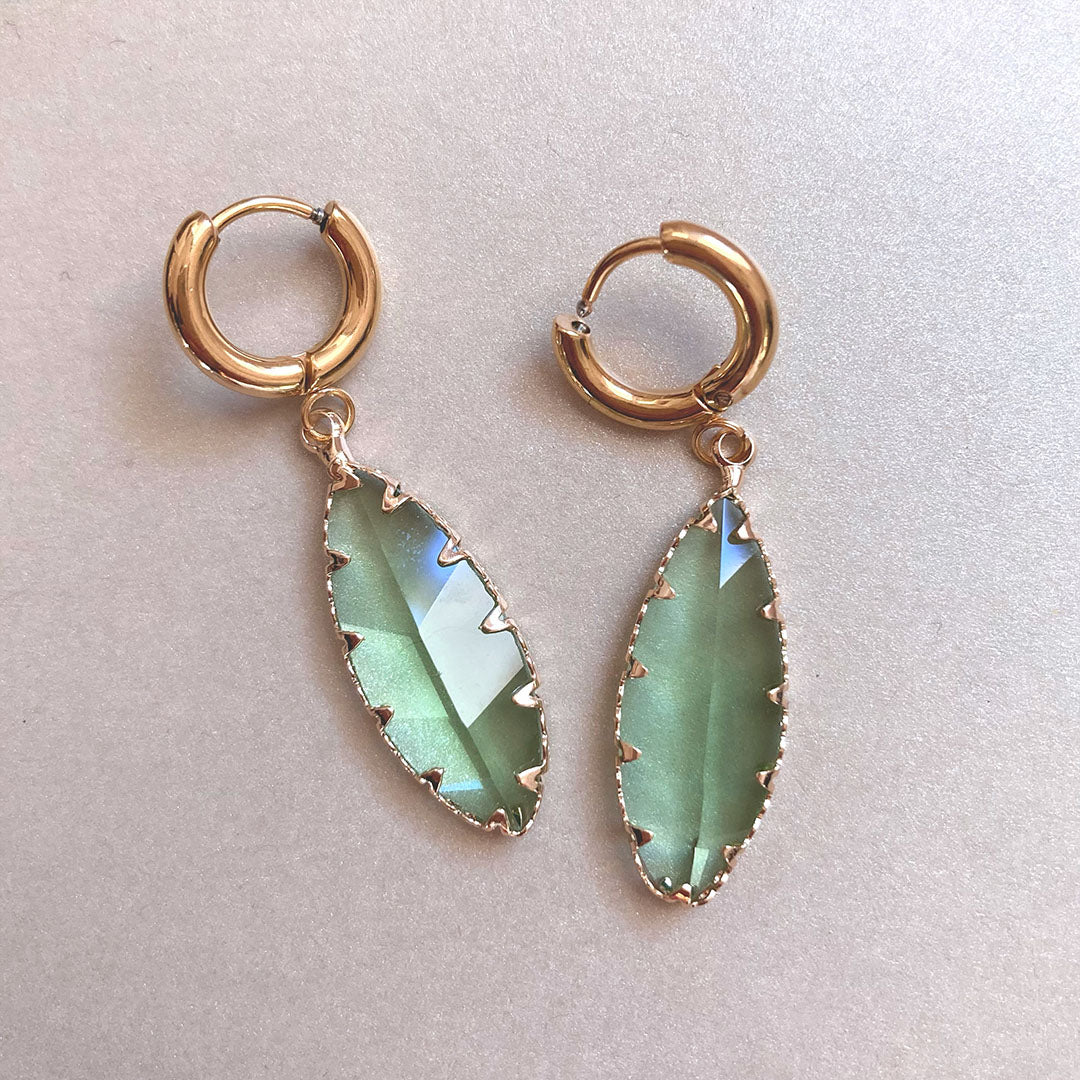 Ein Paar goldfarbene Creolen mit hängenden, blattförmigen Schmucksteinen in Salbeigrün. Die Steine haben einen facettierten Schliff und eine edle Fassung mit kleinen Zacken entlang der Kanten.