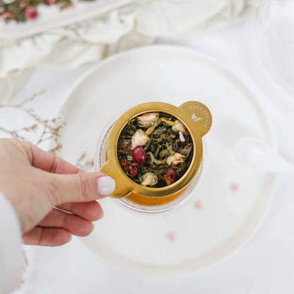 Eine Hand hält ein goldenes Teesieb über einer Tasse, gefüllt mit hochwertigem losem grünem Tee, ganzen Rosenblüten und Beeren.Teller.