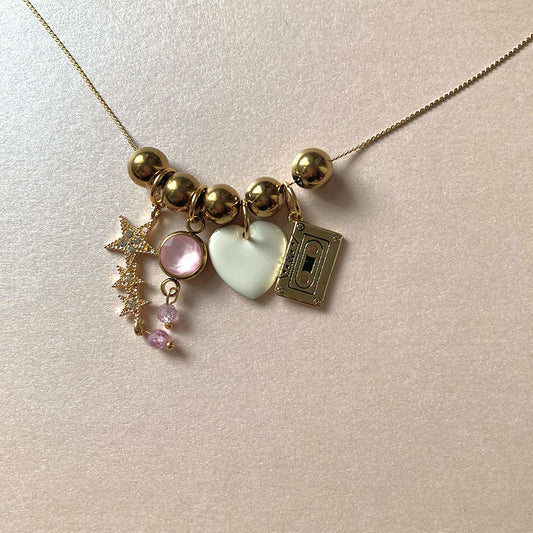 Goldene Halskette mit verschiedenen Anhängern: eine kleine Musikkassette, ein weißes Herz, glitzernde Sterne und rosa Perlen.