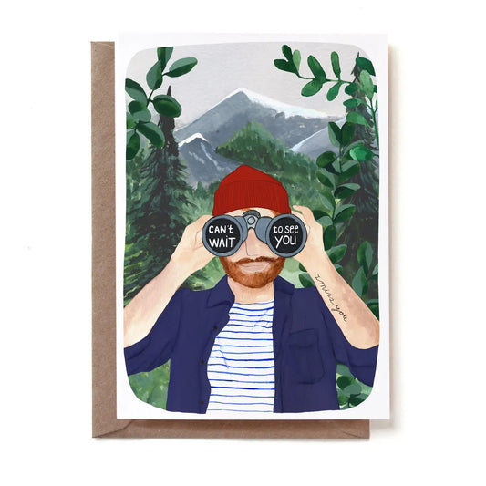 Eine illustrierte Klappkarte mit einem bärtigen Mann, der eine rote Strickmütze, ein blau-weiß gestreiftes T-Shirt und ein offenes blaues Hemd trägt. Er blickt durch ein Fernglas direkt nach vorne. Auf den Linsen des Fernglases steht der weiße Text „CAN'T WAIT to see YOU“. Auf seinem linken Unterarm ist fein „I miss you“ tätowiert. Im Hintergrund erstreckt sich eine Berglandschaft mit Tannen. Die Karte steckt in einem braunen Kraftpapier-Umschlag.