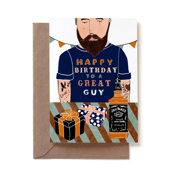 Eine illustrierte Geburtstagskarte für Männer mit einem bärtigen, tätowierten Mann im Comic-Stil, der ein dunkelblaues T-Shirt mit der Aufschrift „HAPPY BIRTHDAY TO A GREAT GUY“ trägt. Im Vordergrund auf einem gestreiften Tisch stehen zwei verpackte Geschenke und eine Flasche Jack Daniel's Whiskey. Die Karte steckt in einem braunen Kraftpapier-Umschlag.
