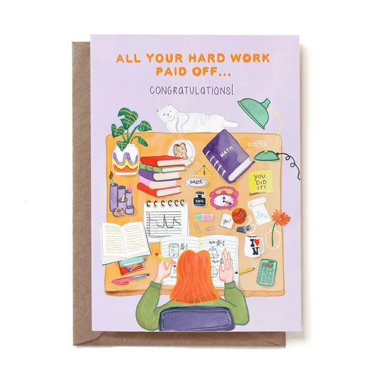 Eine illustrierte Glückwunschkarte zum bestandenen Abschluss mit der Aufschrift „ALL YOUR HARD WORK PAID OFF... CONGRATULATIONS!“. Das Motiv zeigt aus der Vogelperspektive eine Studentin mit roten Haaren am Schreibtisch, die in ihre Mathe-Hefte vertieft ist. Der Tisch ist voll mit Büchern, einem Mathebuch, einem Laptop, Notizen, einem Taschenrechner und einer Kaffeetasse. Eine weiße Katze liegt entspannt am oberen Schreibtischrand. Die Karte steckt in einem braunen Kraftpapier-Umschlag.