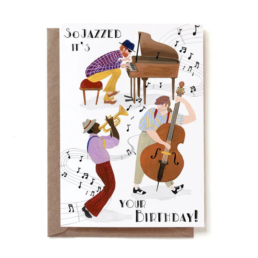 Eine illustrierte Jazz-Geburtstagskarte mit der Aufschrift „So JAZZED IT'S YOUR BIRTHDAY!“. Drei Musiker bilden eine Band: Ein Mann im karierten Hemd spielt Klavier, ein barfüßiger Mann in roter Hose bläst die Trompete und ein Mann mit Schiebermütze spielt Kontrabass. Um sie herum schweben gezeichnete Notenlinien und Musiknoten auf weißem Grund. Die Karte steckt in einem braunen Kraftpapier-Umschlag.