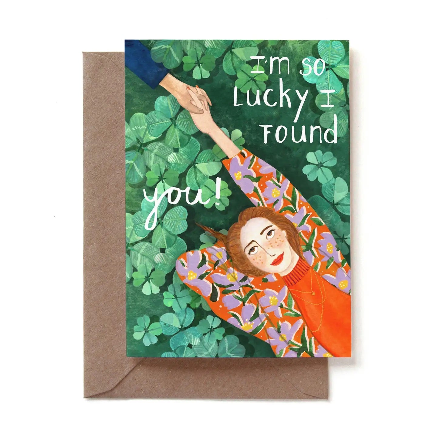 Eine liebevoll illustrierte Karte mit der weißen Aufschrift „I'm so lucky I found you!“. Das Motiv zeigt eine Frau mit Sommersprossen und rotbraunem Haar aus der Vogelperspektive, die rücklings in einer dichten Wiese aus vierblättrigen Kleeblättern liegt. Sie trägt eine auffällige orangefarbene Jacke mit lila Blumenmuster und streckt den Arm nach oben, um die Hand einer anderen Person zu halten, die von außerhalb des Bildrandes ins Motiv reicht. Die Karte steckt in einem braunen Kraftpapier-Umschlag.