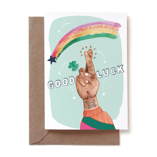 Eine illustrierte Glückwunschkarte mit der Aufschrift „GOOD LUCK“. Das Motiv zeigt eine Hand mit tätowiertem Handgelenk (ein Hufeisen mit Herz), die die Finger als Glückssymbol kreuzt und ein kleines vierblättriges Kleeblatt hält. Über der Hand spannt sich ein bunter Aquarell-Regenbogen mit dem Text „WISHING YOU THE BEST OF LUCK!“. Der Hintergrund ist in hellem Mintgrün gehalten. Die Karte steckt in einem braunen Kraftpapier-Umschlag.