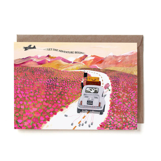 Eine illustrierte Hochzeitskarte im Querformat. Ein weißer Oldtimer-Käfer mit zwei Koffern auf dem Dach und einem „JUST MARRIED“-Nummernschild fährt eine geschwungene Straße entlang, während Blechdosen an Schnüren hinterhergezogen werden. Das Auto fährt durch ein riesiges, leuchtend pinkes Feld voller Blumen auf herbstliche, rötliche Berge zu. Am Himmel fliegt ein kleines Flugzeug, das ein Banner mit der Aufschrift „LET THE ADVENTURE BEGIN!“ hinter sich herzieht. Die Karte liegt auf einem braunen Kraftpapie