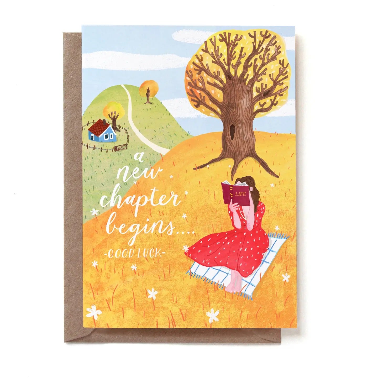 Eine illustrierte Glückwunschkarte mit der Aufschrift „a new chapter begins... -GOOD LUCK-“. Auf einer herbstlich gelben Wiese sitzt eine Frau in einem roten Kleid auf einer Picknickdecke und liest in einem Buch mit dem Titel „LIFE“. Sie sitzt im Schatten eines großen, knorrigen Baumes. Im Hintergrund führt ein Weg einen grünen Hügel hinauf zu einem kleinen blauen Haus. Die Karte steckt in einem braunen Kraftpapier-Umschlag.