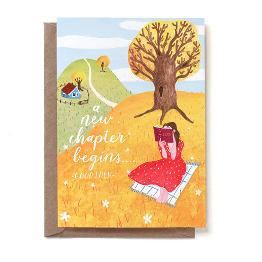 Eine illustrierte Glückwunschkarte mit der Aufschrift „a new chapter begins... -GOOD LUCK-“. Auf einer herbstlich gelben Wiese sitzt eine Frau in einem roten Kleid auf einer Picknickdecke und liest in einem Buch mit dem Titel „LIFE“. Sie sitzt im Schatten eines großen, knorrigen Baumes. Im Hintergrund führt ein Weg einen grünen Hügel hinauf zu einem kleinen blauen Haus. Die Karte steckt in einem braunen Kraftpapier-Umschlag.