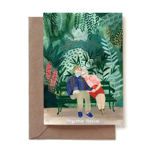 Eine illustrierte Grußkarte mit einem älteren Paar, das glücklich Hand in Hand auf einer grünen Parkbank sitzt. Der Mann trägt ein blau kariertes Hemd mit Hosenträgern, die Frau ein rosa Oberteil und einen roten Rock. Sie sind von einem dichten, grünen Garten mit Farnen und roten Blumen umgeben. Am unteren Rand steht der Text „Together Forever“. Die Karte steckt in einem braunen Kraftpapier-Umschlag.