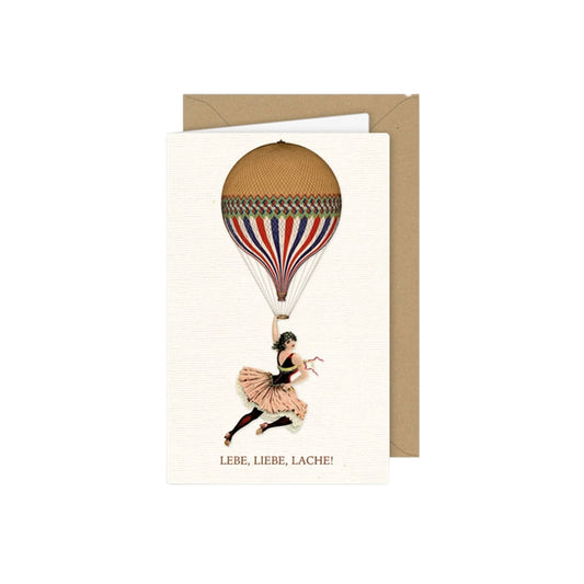 Vintage-Grußkarte mit einer Akrobatin/Tänzerin im Rosa Tüllkleid, die sich an der Halterung eines gestreiften Heißluftballons in Rot, Blau, Weiß und Gold festhält. Darunter steht der Text „LEBE, LIEBE, LACHE!“.