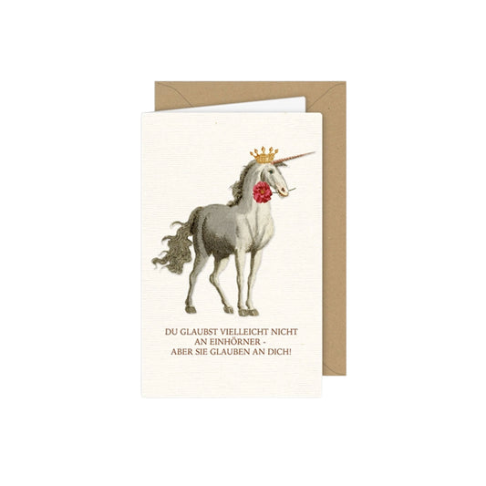 Magische Grußkarte mit einem eleganten grauen Einhorn mit Goldkrone, das eine rote Blume im Maul hält. Darunter steht der spruch: „DU GLAUBST VIELLEICHT NICHT AN EINHÖRNER - ABER SIE GLAUBEN AN DICH!“.